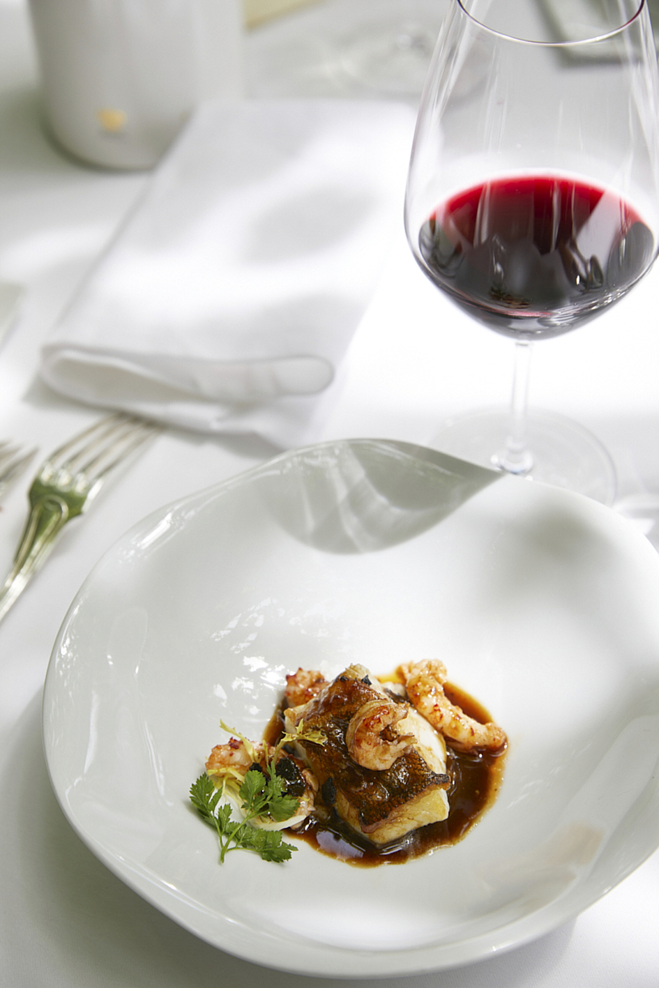 Photographie professionnelle d'un plat gastronomique de poisson et crevettes en sauce, servi dans une assiette design avec un verre de vin rouge. Image réalisée par Studio ZE, photographe culinaire de haute cuisine à Toulouse.