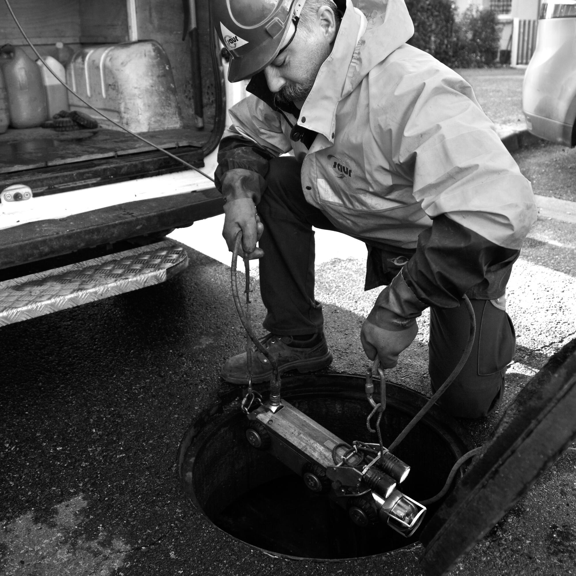 Photographie en noir et blanc d'un homme en tenue de travail inspectant un conduit souterrain avec un équipement spécifique. Par Studio ZE, photographe humain à Toulouse.
