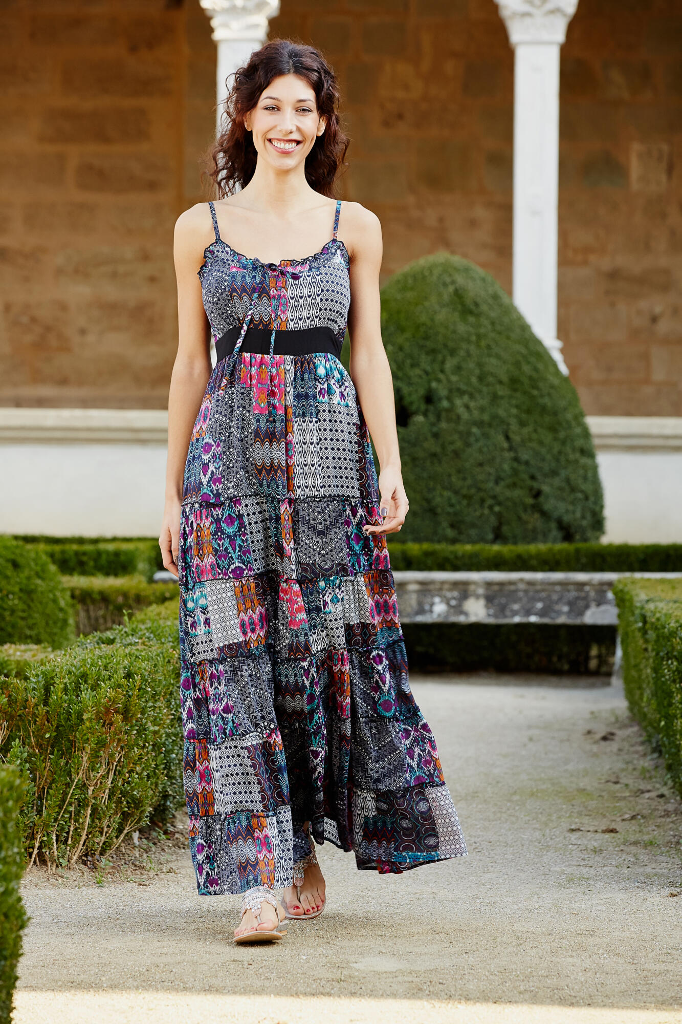 Photographie d'une femme souriante aux cheveux bruns, vêtue d'une robe longue à motifs colorés, marchant dans un jardin. Par Studio ZE, photographe mode à Toulouse.
