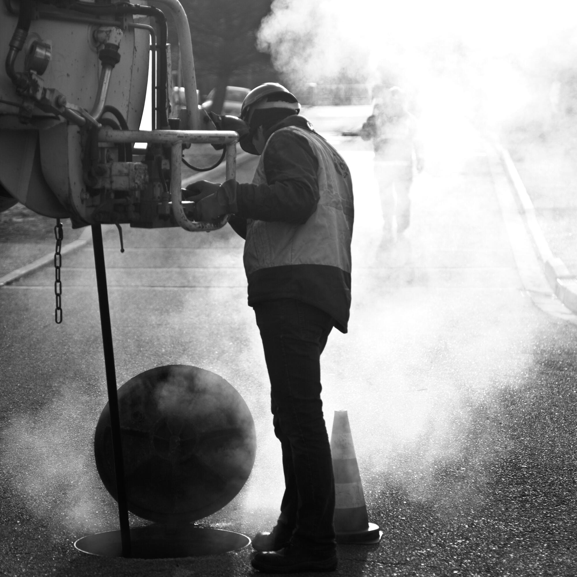 Photographie en noir et blanc d'un ouvrier en silhouette avec casque, travaillant dans la vapeur d'une bouche d'égout. Par Studio ZE, photographe humain à Toulouse.