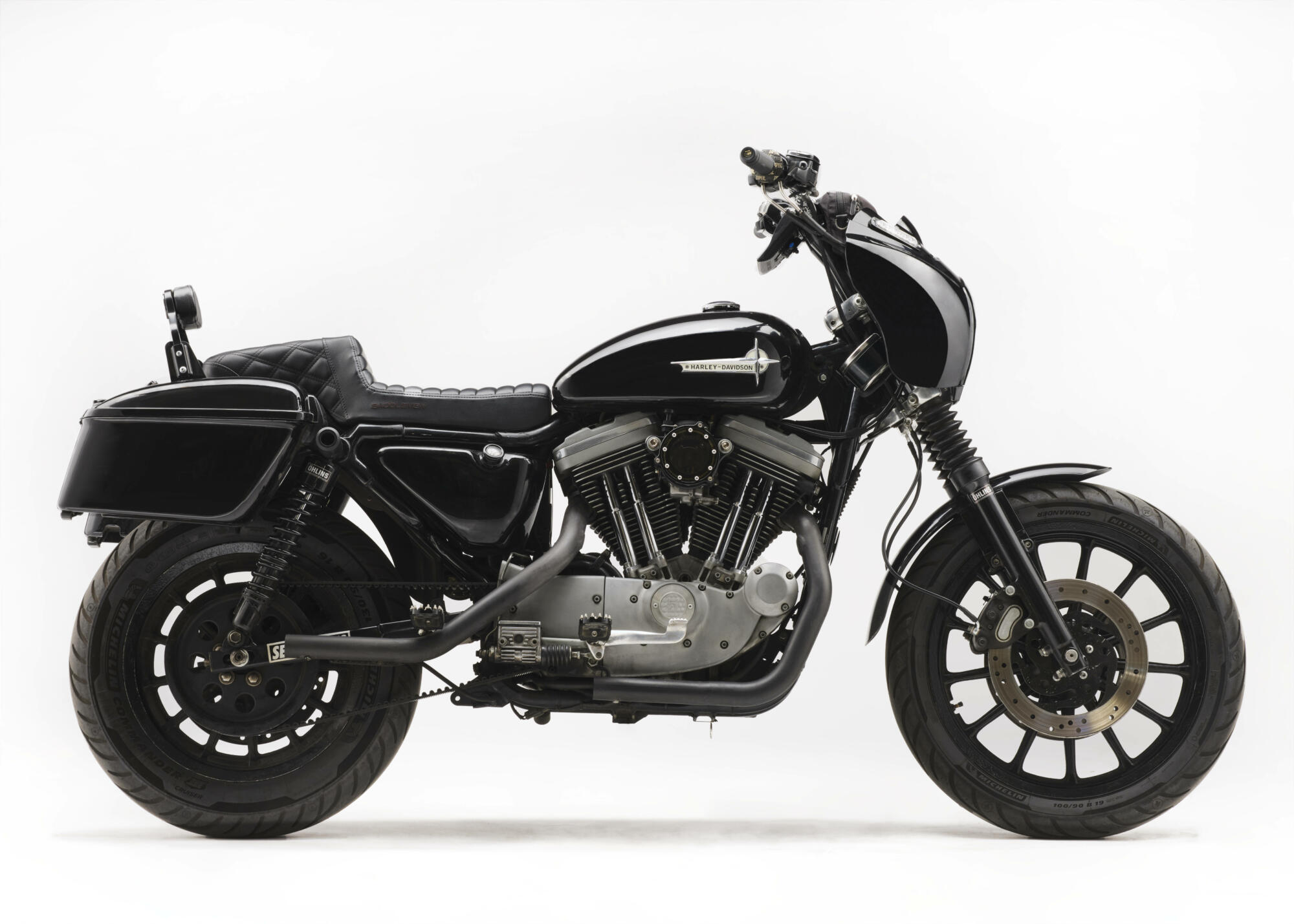 Photographie d'une moto Harley-Davidson Sportster noire customisée, avec des sacoches latérales et des échappements apparents, sur fond blanc. Par Studio ZE, photographe auto moto à Toulouse.