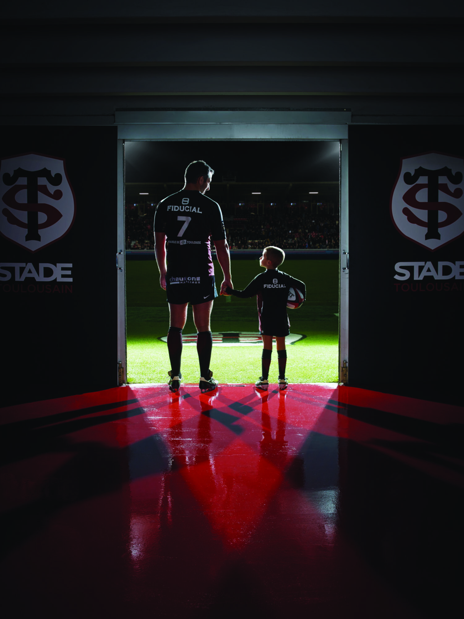 Photographie d'un joueur de rugby du Stade Toulousain tenant la main à un enfant en se regardant, ambiance avant match, entrée dans le stade. Par Studio ZE, photographe humain à Toulouse.