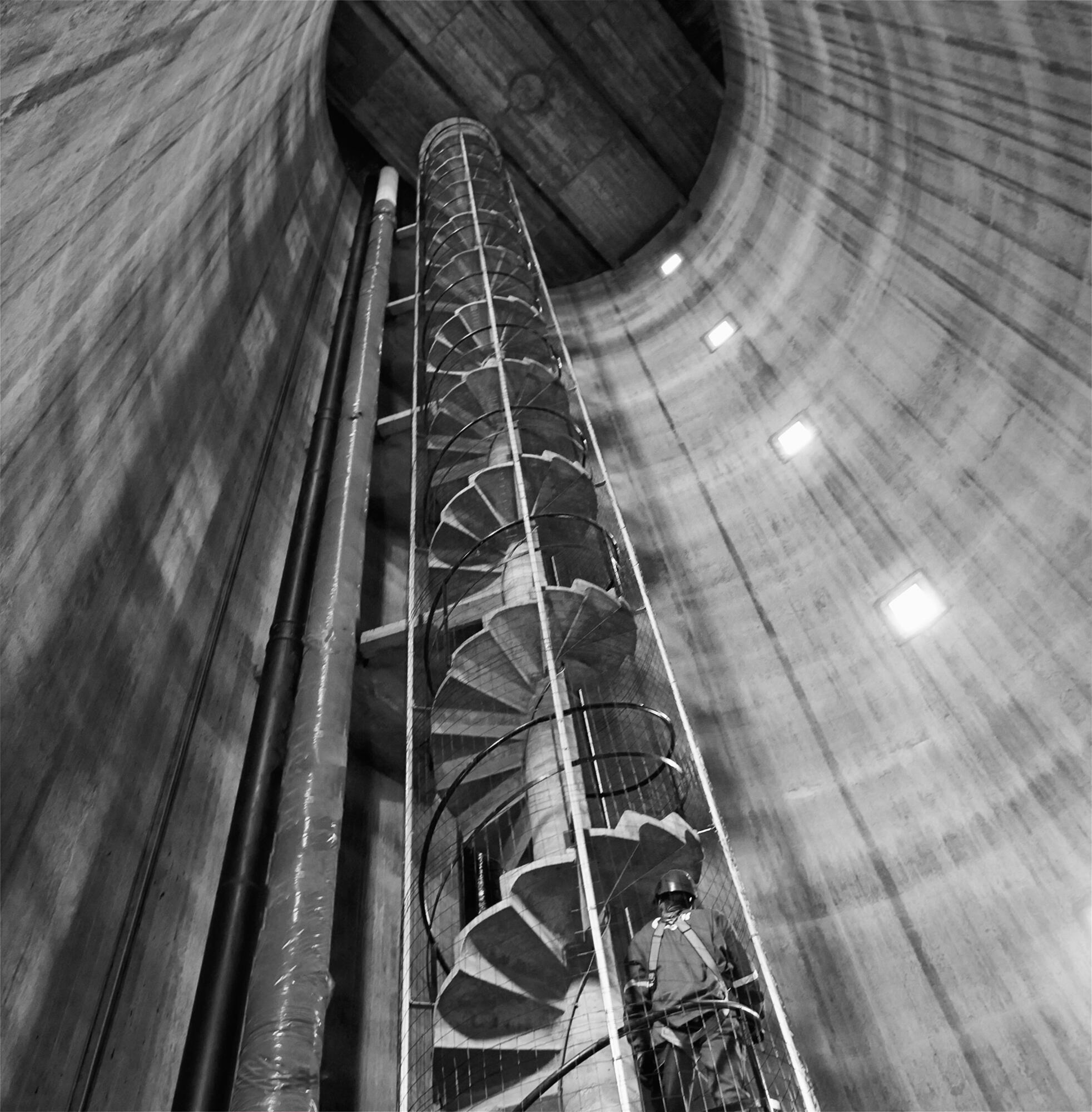 Photographie en noir et blanc d'un ouvrier casqué dans une structure industrielle cylindrique avec escalier en spirale. Par Studio ZE, photographe humain à Toulouse.