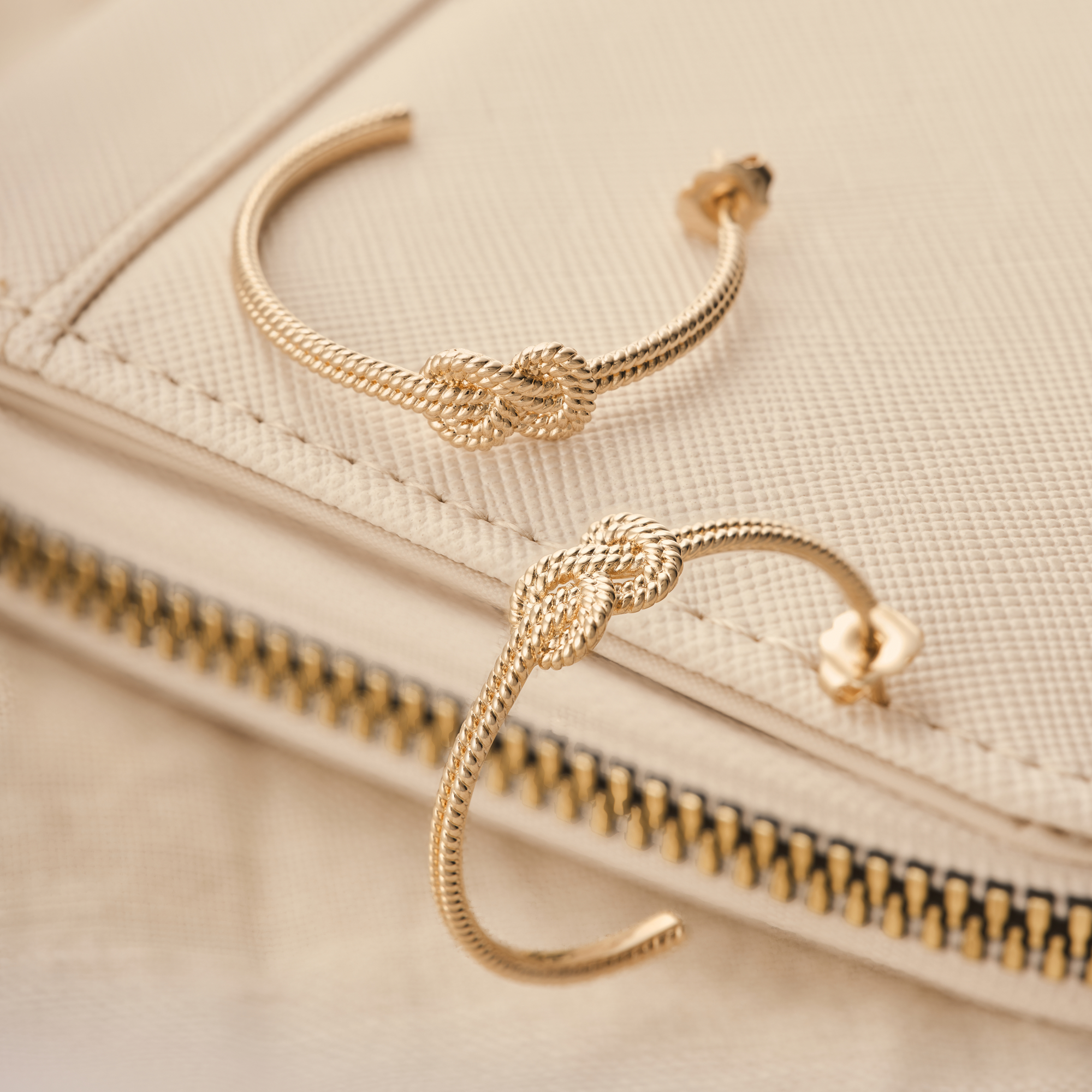 Photographie de boucles d'oreilles créoles dorées avec un détail de nœud, posées sur un sac en cuir clair. Par Studio ZE, photographe bijoux à Toulouse.