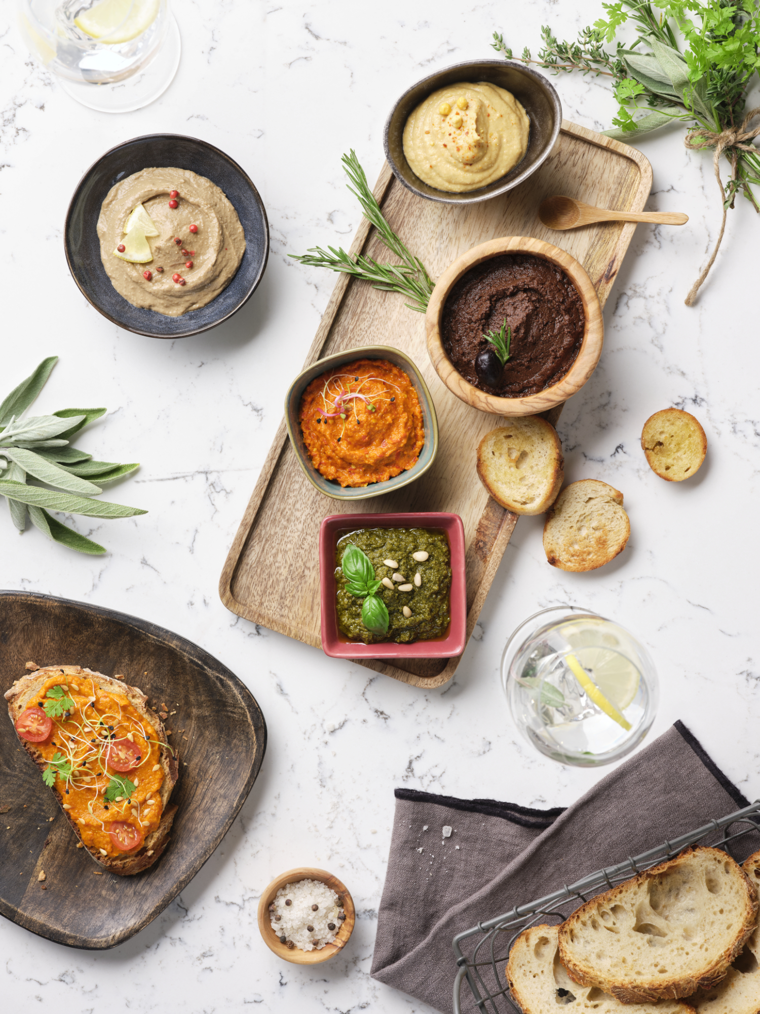 Photographie de divers dips et tartinades (houmous, pesto, tapenade) avec pain grillé et herbes. Par Studio ZE, photographe culinaire à Toulouse.