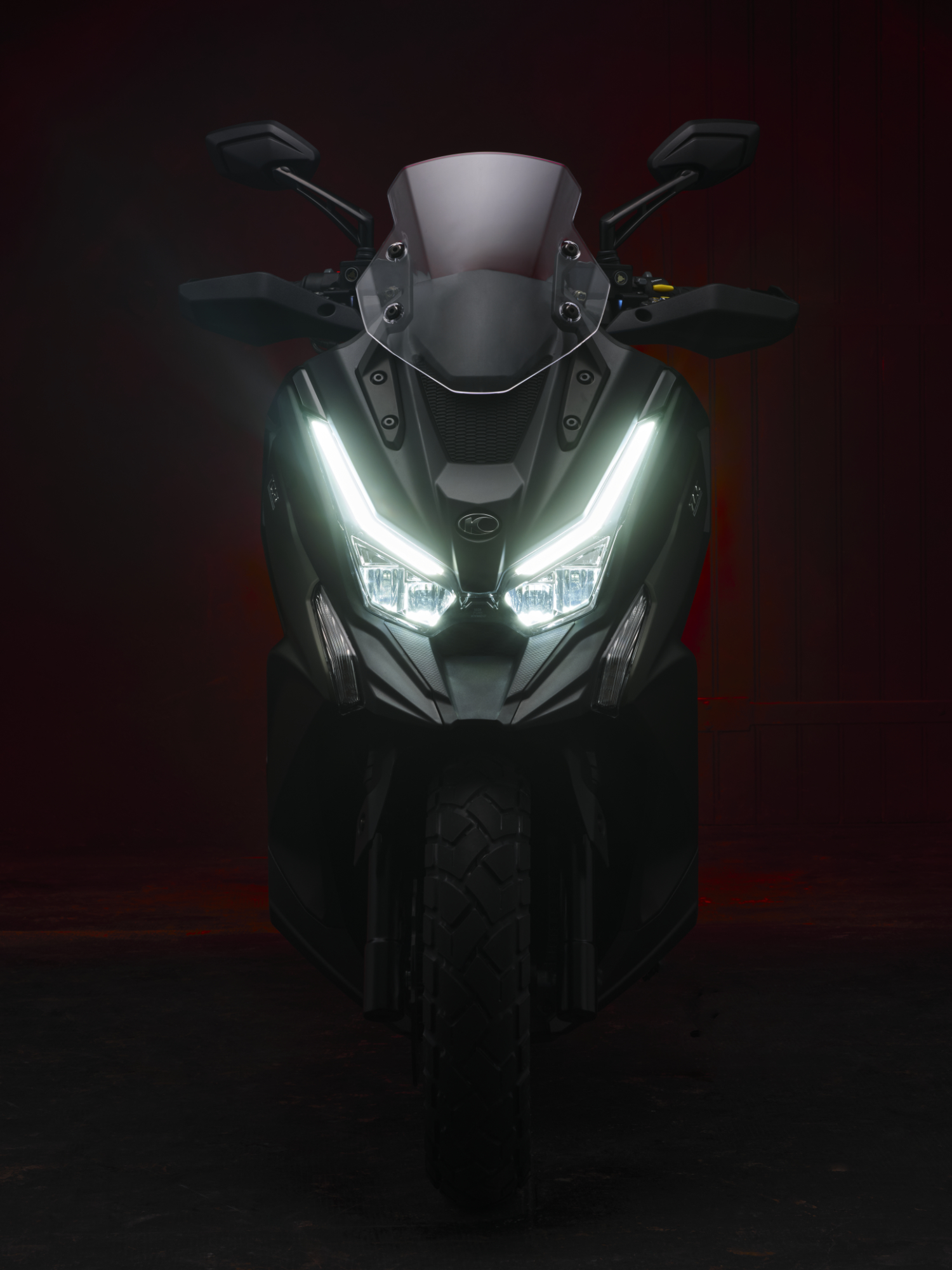 Photographie de face d'un scooter sombre avec des phares LED distinctifs allumés, sur fond rouge foncé. Par Studio ZE, photographe auto moto à Toulouse.