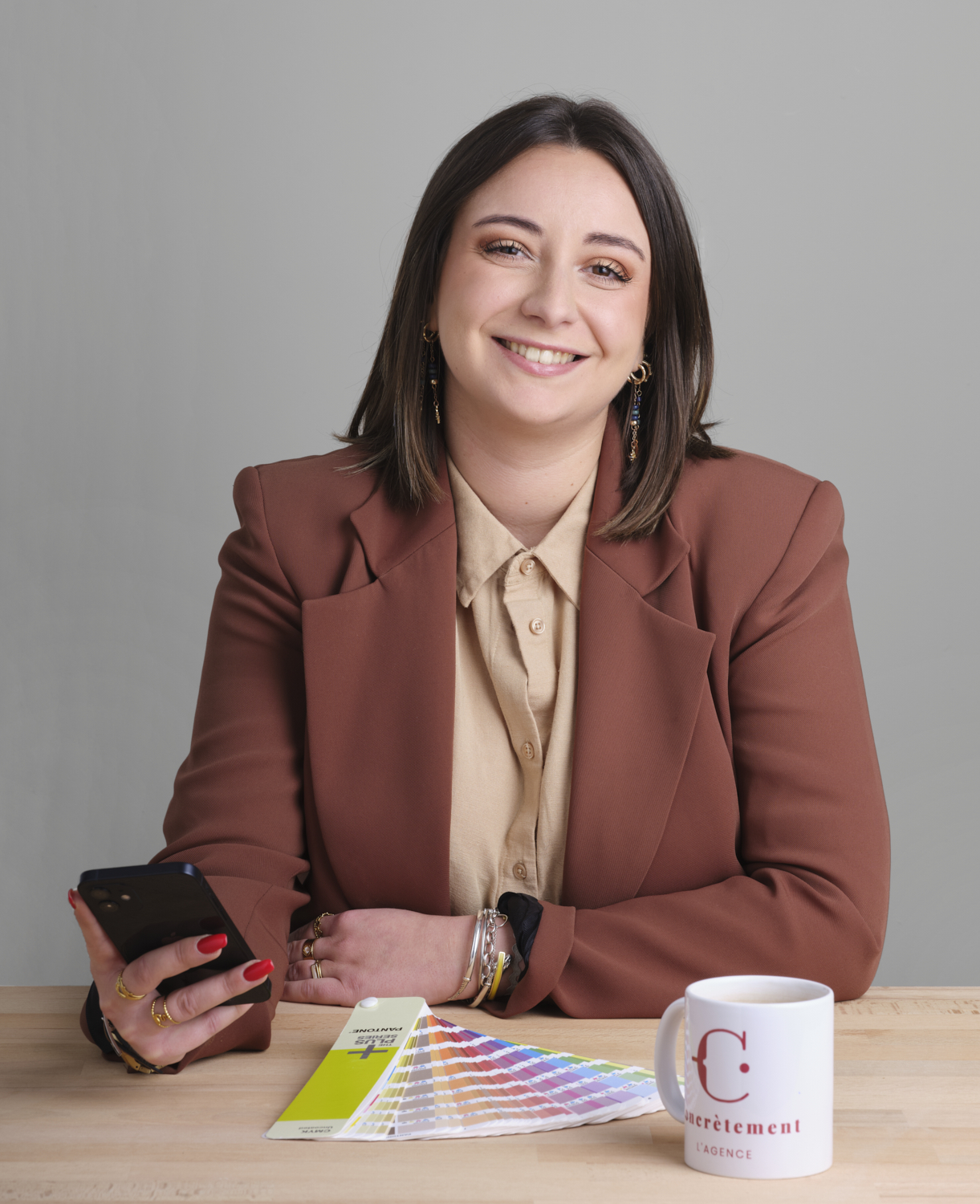 Photographie d'une femme souriante assise à un bureau avec un smartphone, une tasse et un nuancier. Par Studio ZE, photographe humain à Toulouse.