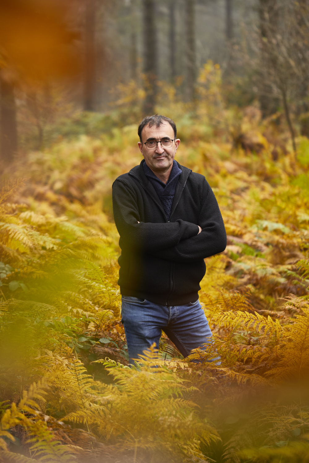 Photographie d'un homme en veste noire et jean, les bras croisés, dans une forêt aux couleurs automnales. Par Studio ZE, photographe humain à Toulouse.