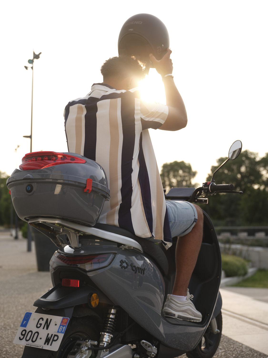 Photographie d'un homme en chemise rayée et short, mettant son casque sur un scooter, avec un coucher de soleil en arrière-plan. Par Studio ZE, photographe humain à Toulouse.